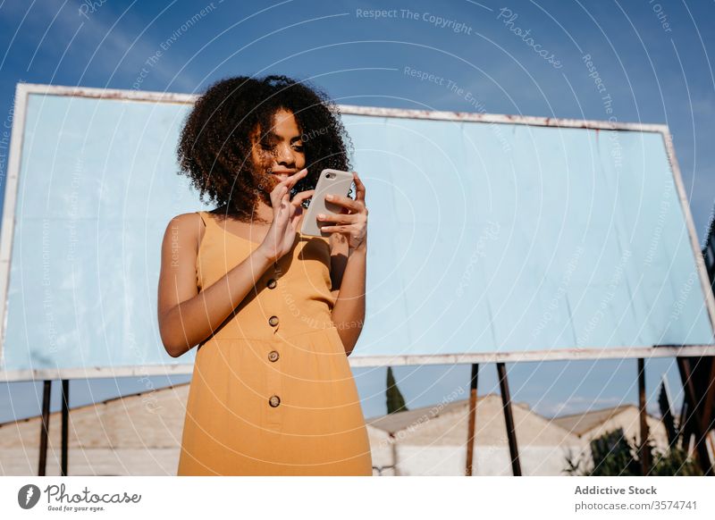 Junge zufriedene ethnische Frau benutzt Handy auf der Straße gegen leere große Tafel in einem Vorort Smartphone Plakatwand verwenden Freude Browsen Surfen lesen