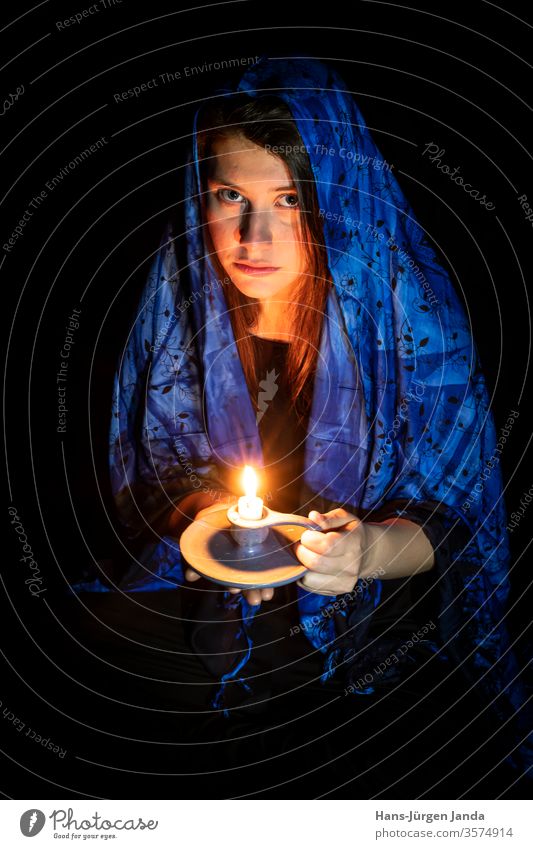 Traurige junge Frau mit Kerze und blauem Kopftuch vor schwarzem Hintergrund Gesicht frau traurig kopftuch portrait frontal profil ernst young kunst düster