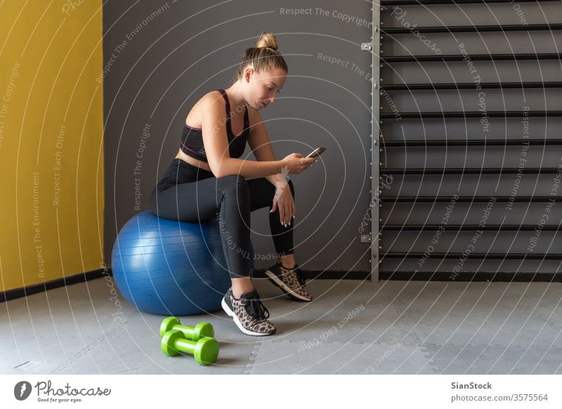 Sportlerin, die nach dem Training oder der Übung im Fitnessstudio sitzt, sich ausruht und ihr Handy beobachtet. App Mobile Telefon sportlich schlank Pause