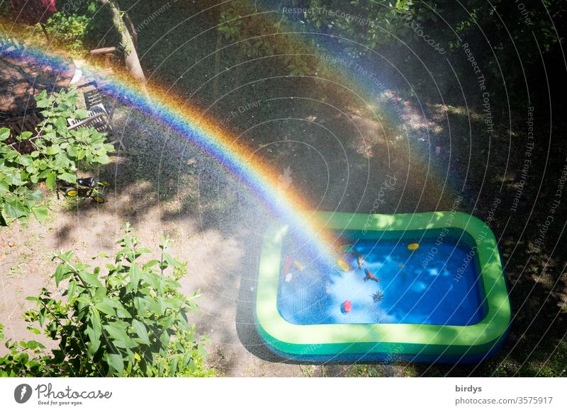 Regenbogen über einem Kinderplanschbecken in einem Garten Planschbecken Wasser Spielzeug Wassertropfen sprühnebel Sonnenlicht Kindheit draußen Schönes Wetter