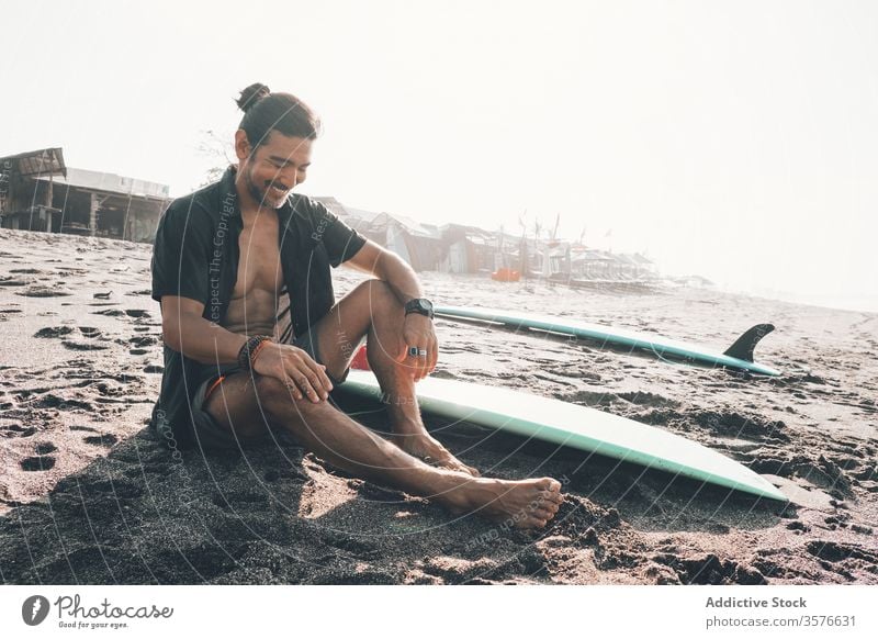 Glücklicher ethnischer Mann mit Surfbrett am Strand sitzend positiv ruhen MEER genießen Sand Sommer jung hispanisch männlich Lifestyle Urlaub Küste Meer Ufer