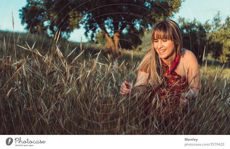 Hübsches junges blondes Modell, das bei Sonnenuntergang in einem Feld lächelt und ein Kleid trägt. Glückliche freie Frau in der Natur. Gemütliche Fotografie.