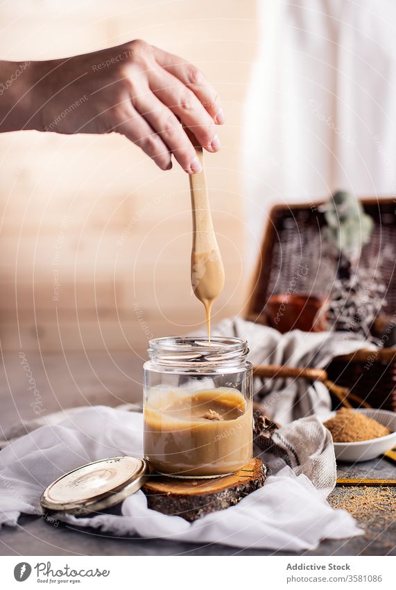 Nutzpflanzenfrau bereitet Karamell bei Tisch in einer hellen, modernen Küche zu Glas Kaffeelöffel Hand Frau Zucker Gewebe selbstgemacht Dessert Leckerbissen