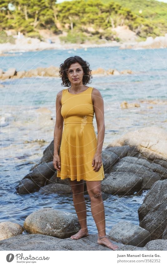 Eine schöne weibliche Latina in einem gelben Kleid steht auf einem Felsen am Meeresufer Frau Mädchen Schönheit jung hübsch attraktiv Menschen Erwachsener weiß