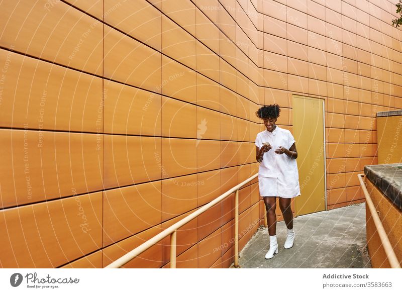 Glückliche schwarze Frau mit Smartphone auf der Straße gegen lebhaftes Äußeres eines modernen Gebäudes Nachrichtenübermittlung Weg Kälte Browsen verwenden