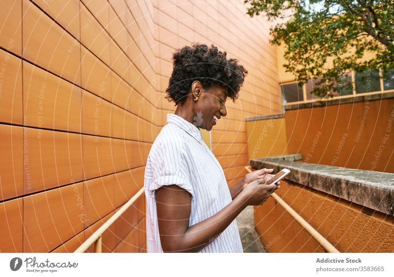 Glückliche schwarze Frau mit Smartphone auf der Straße gegen lebhaftes Äußeres eines modernen Gebäudes Nachrichtenübermittlung Weg Kälte Browsen verwenden