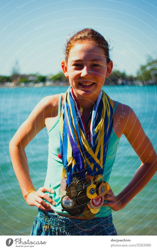 Mein Champion.//  Ein Mädchen steht fröhlich am Uferrand , lachend. Sie hat ganz viele Medallien um den Hals gehängt . Sie hat die Arme angewinckelt. girl