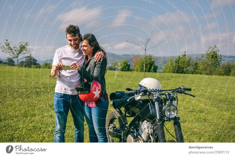 Paar, das mit einem Motorrad mobil aussieht Glück Blick Smartphone Bild Video Ausflug Mann Lächeln Handy umarmend Motorradhelm benutzerdefiniert attraktiv