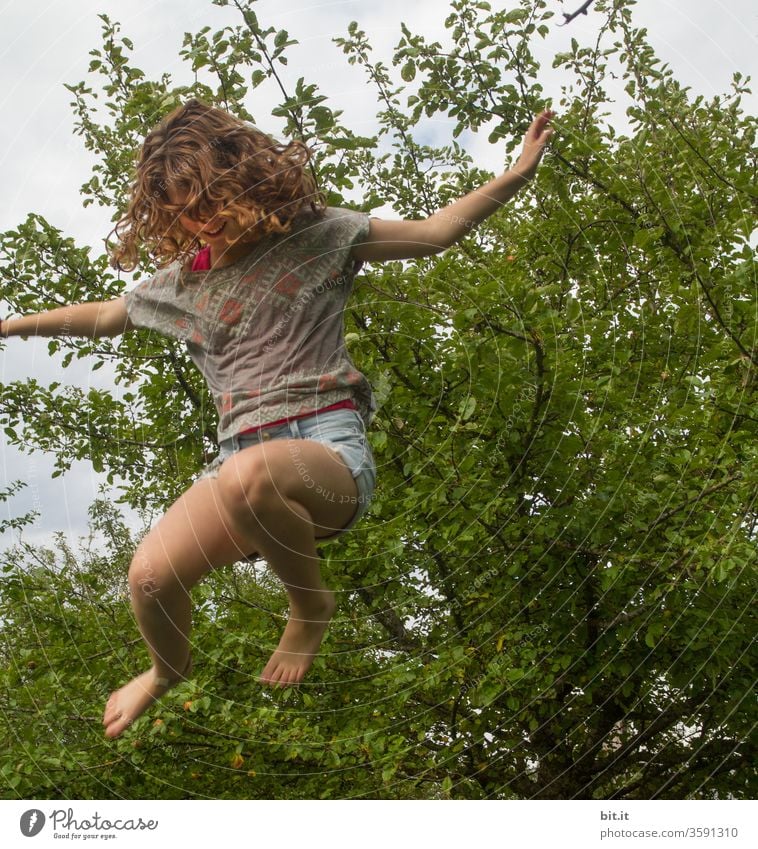 Lockiger, temperamentvoller Spaßvogel springt auf Trampolin im Garten. Mädchen fliegt, hüpft, springt lebensfroh, glücklich, wild, verrückt nach oben. Sport, Unbeschwertheit, Bewegung und Fitness gegen Stress, Probleme, Angst in der Schule und zu Hause.
