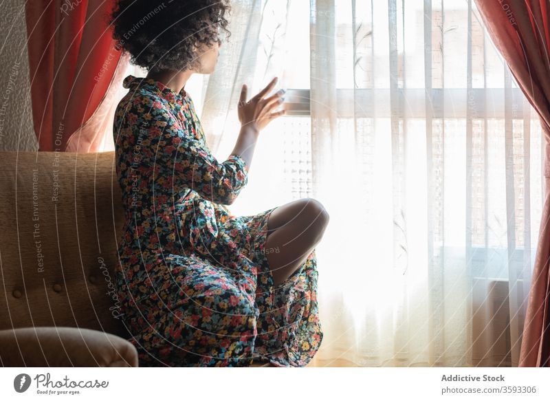 Nachdenkliche schwarze Frau im Kleid am Fenster im Wohnzimmer besinnlich ruhen Armsessel Windstille sich[Akk] entspannen friedlich Stil ethnisch Afroamerikaner