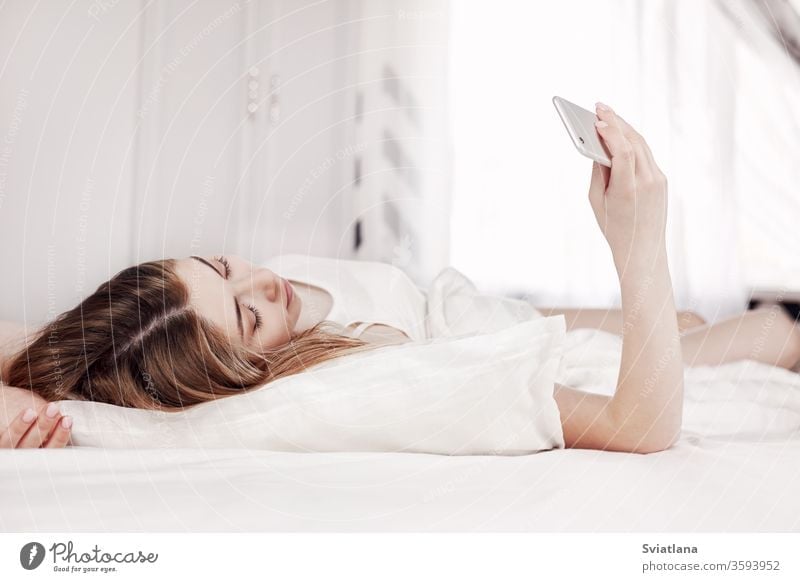 Ein hübsches junges Mädchen schaut auf das Telefon, während sie auf dem Bett liegt. Ein Mädchen liest Nachrichten auf ihrem Telefon, wenn sie morgens aufwacht
