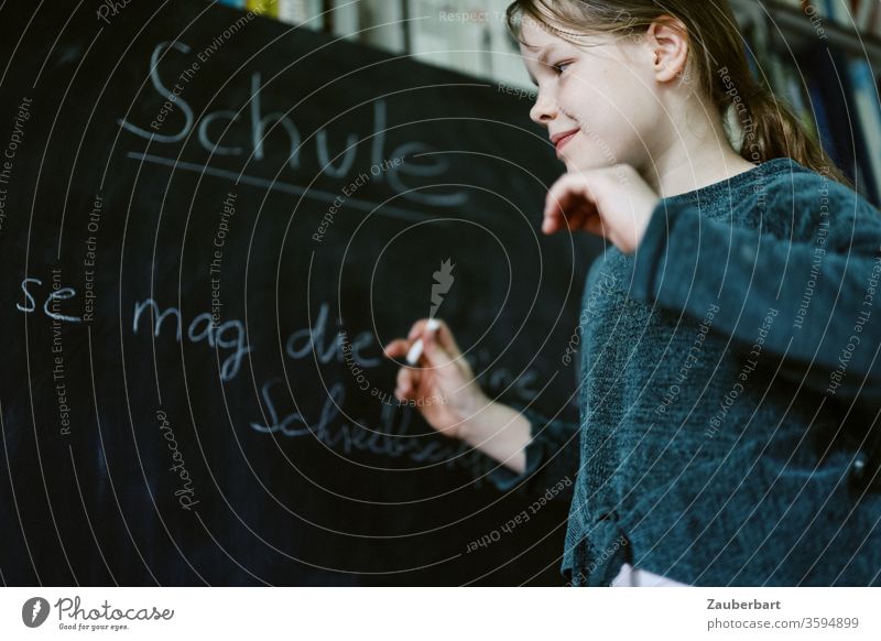 Homeschooling VII - Mädchen spielt Schule und schreibt mit Kreide auf eine Tafel einen Übungssatz für Schreibschrift, im Hintergrund Bücherregal Schülerin