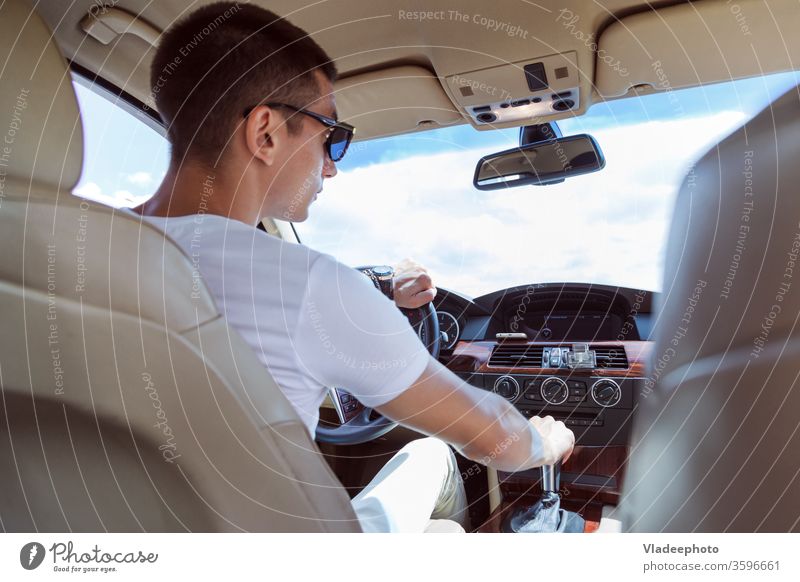Junger stilvoller Mann mit Sonnenbrille, der ein Auto fährt. Blick von hinten, mit dem Beifahrersitz im Fond. PKW männlich Fahrzeug Automobil im Inneren Straße