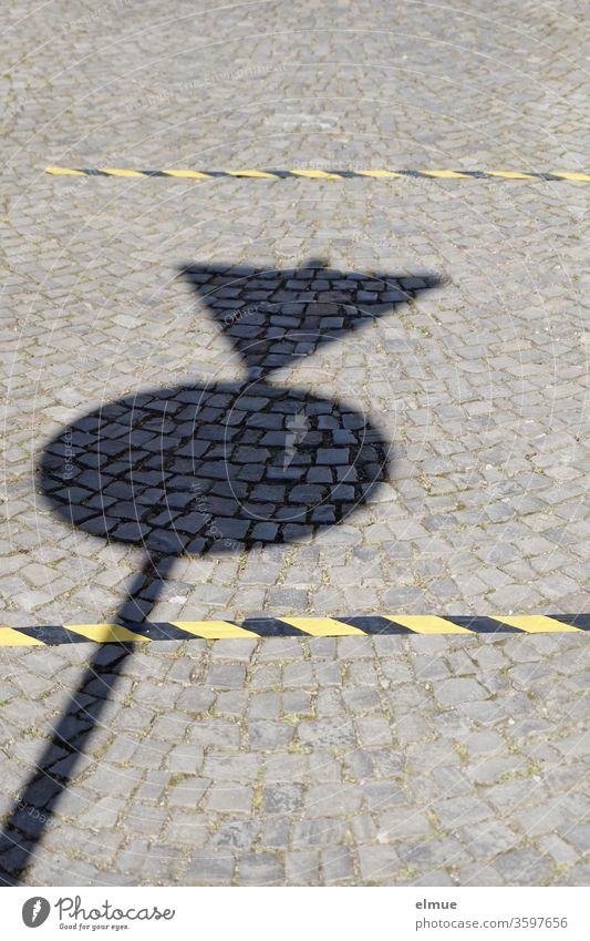 Schatten eines Verkehrsschildes auf einem gepflasterten Weg und zwei schwarz-gelbe Markierungen zur Abstandshaltung Abstand halten Infektionsschutz