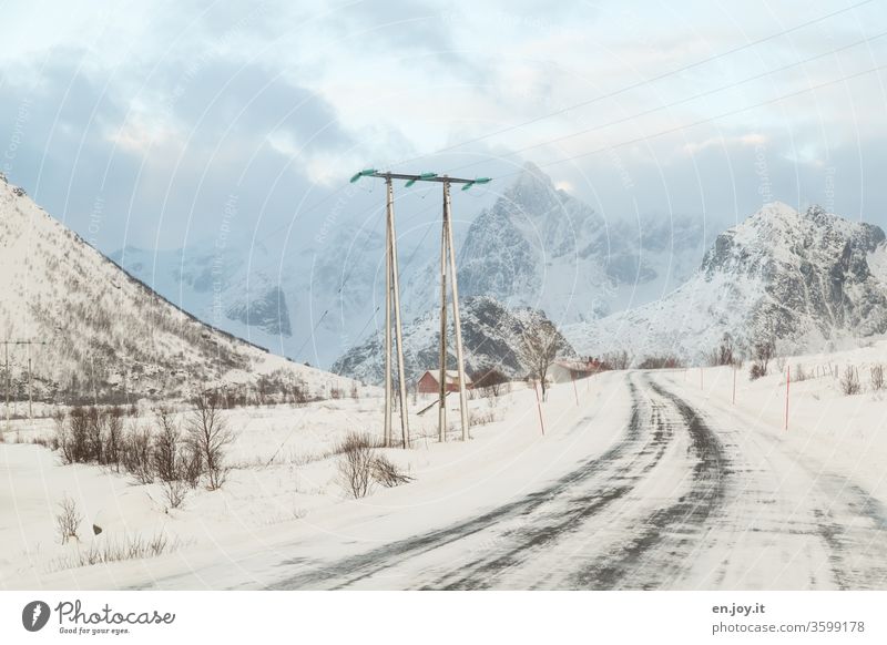 verschneite Straße in Norwegen führt unter einer Stromleitung hindurch vor Bergen mit Schnee Lofoten Skandinavien Winter Strommast Energieversorgung