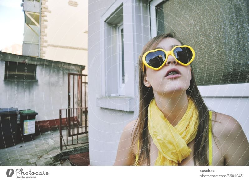 jugendliche mit gelber herzsonnenbrille und gelbem schal in gelbem bikini steht in einem etwas heruntergekommenen hinterhof und schaut nach oben Mädchen