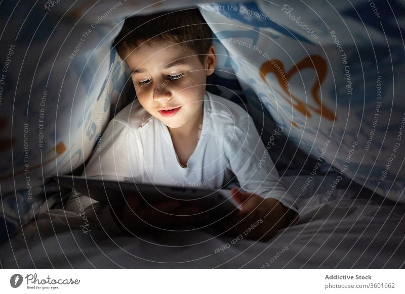 Entzückter Junge sieht Zeichentrickfilm auf dem Tablett Kind benutzend Tablette Decke Nacht zuschauend heiter Karikatur Pyjama heimwärts bezaubernd Gerät