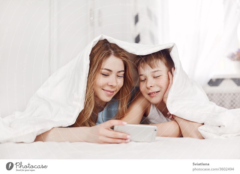 Ein hübsches junges Mädchen und ihr fröhlicher jüngerer Bruder sehen sich Fotos auf ihrem Telefon an, während sie lächelnd auf dem Bett liegen. Bruder und Schwester schauen sich Videos auf ihrem Telefon an.