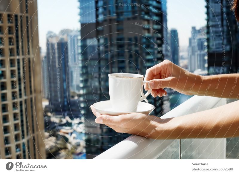 Ein Mädchen hält eine Tasse Kaffee vor dem Hintergrund moderner Gebäude. Eine Frau trinkt während der Selbstisolation auf dem Balkon einen Morgenkaffee