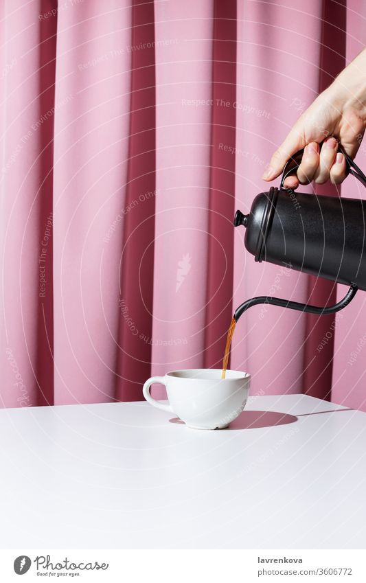 Frauenhand hält schwarze Metallkanne und gießt Kaffee oder Tee in weiße Tasse vor rosa Draperie, selektiver Fokus Getränk Morgen trinken Becher Frühstück