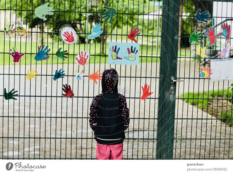 Ein Kind in Regenkleidung steht traurig vor  einem geschlossenen Kindergarten.Rückansicht.  Ausgangsbeschränkungen, Corona, Schließungen sehnsucht kindergarten