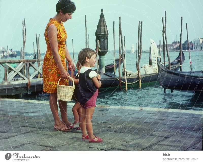 alt l Hübsche, junge, modische Mutter mit zwei Töchtern im Sommerurlaub, stehen im Hafen von Venedig. Mutter mit Kindern beobachtet neugierig Schiffe und Boote im Meer. Familienausflug in Italien in den 60er Jahren. Nostalgisches, altes 60er Familienphoto