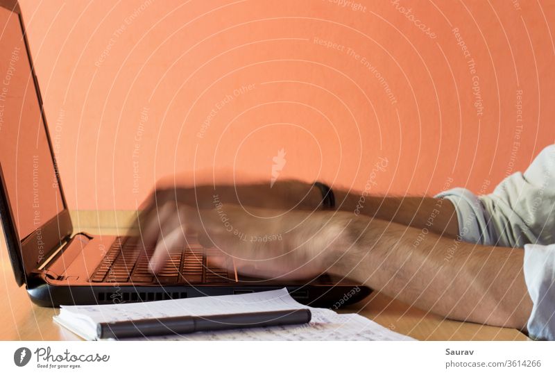 Eine bewegungsunscharfe Nahaufnahme der Finger eines jungen Erwachsenen, der von zu Hause aus auf einem Laptop arbeitet, der auf einem Holztisch neben einem Stift und einem Notizblock aufbewahrt wird.