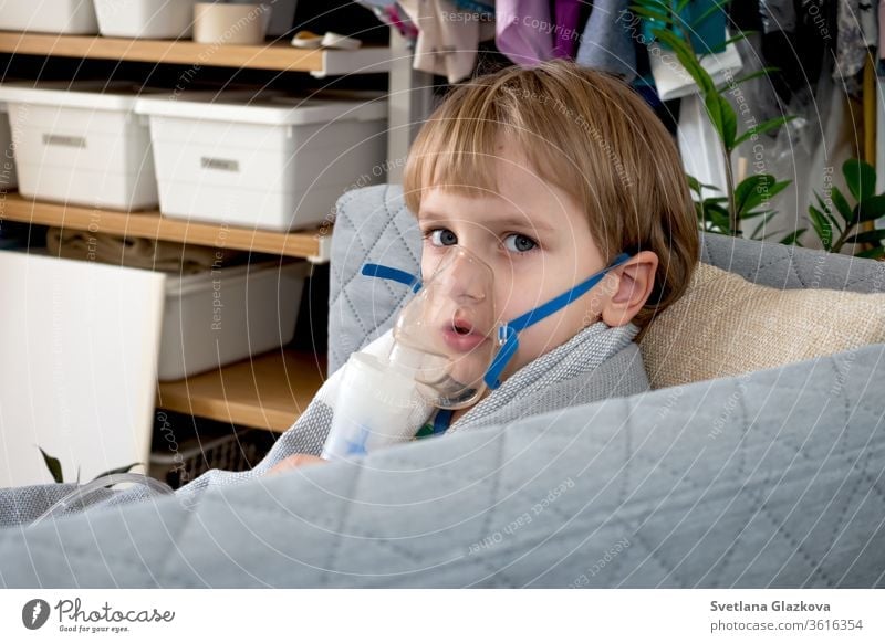 Kleiner kaukasischer Junge, der zu Hause mit einem Vernebler inhaliert. Kind hält einen Maskendampf-Inhalator in der Hand. Behandlung von Asthma. Konzept eines Inhalationstherapiegerätes.