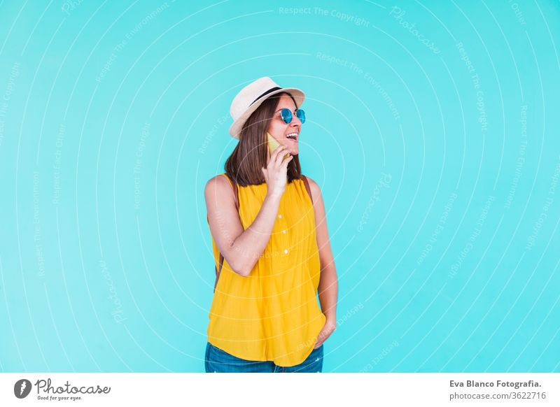 junge Frau, die im Freien vor einem türkisfarbenen Hintergrund mobil telefoniert. Sommerzeit Handy Türkisfarbener Hintergrund Sonnenbrille gelb Hut Rucksack