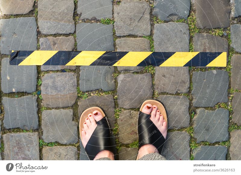 Abstand halten! - weibliche Füße stehen vor einem schwarz-gelben Klebeband auf Kopfsteinpflaster Corona Corona-Virus Schutz Pandemie Infektionsgefahr Gesundheit