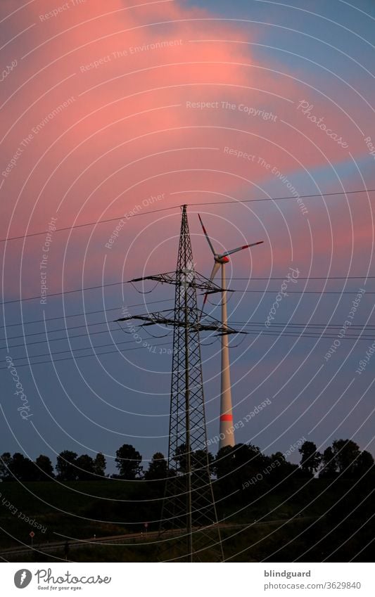 Energiemix. Windrad hinter einem Mast der Überlandleitung für die bundesweite Stromversorgung, mit Wald vor einem abendlich rot-blauen Himmel. Energiewirtschaft