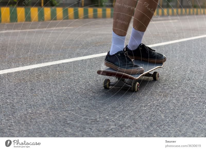Nahaufnahme der Füße eines jungen Teenagers mit zerrissenen schwarzen Turnschuhen auf seinem Skateboard, der auf einer leeren Straße fährt, während die weiße Linie auf der Straße seine weißen Socken ergänzt und auch als Führungslinie in der Richtung dient, in die er sich bewegt.