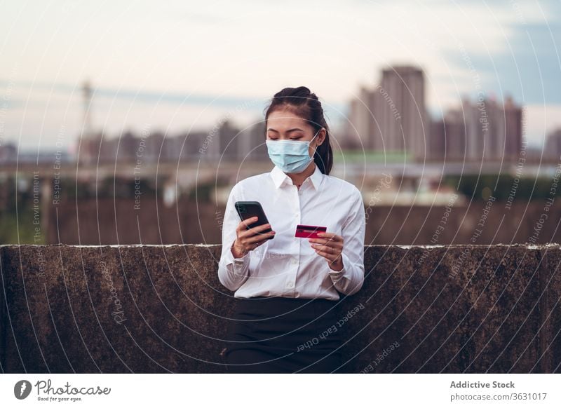 Vielbeschäftigte Frau mit Schutzmaske beim Online-Kauf mit Smartphone Kreditkarte benutzend bezahlen online formal jung Mundschutz Dachterrasse urban asiatisch