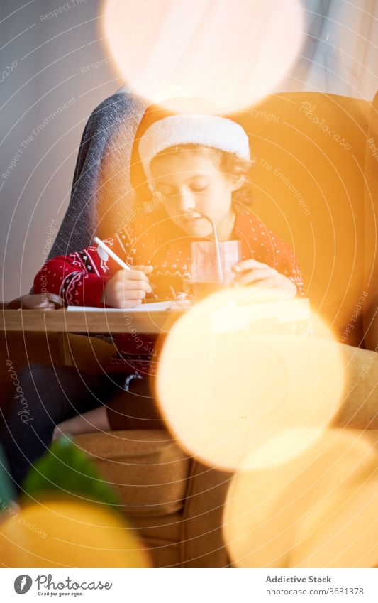 Fröhlicher Junge in Weihnachtskleidung im gemütlichen Sessel sitzend Weihnachtsmütze Weihnachten Feiertag Bleistift Armsessel Glück Komfort heimisch Pullover