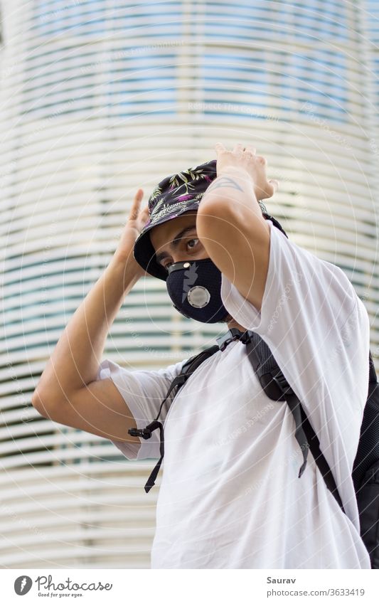 Ein junger Mann im Freien mit einer schützenden Gesichtsmaske zur Verhinderung einer Coronavirusinfektion, der während einer globalen Pandemie seine blumig bedruckte Eimerkappe anpasst und einen Beutelpack trägt.