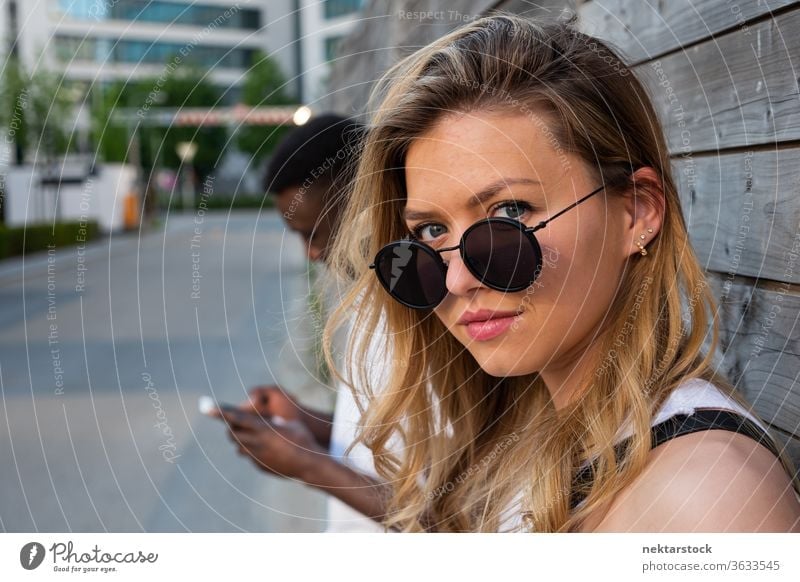 Junge Frau mit runder Sonnenbrille schaut in die Kamera Porträt verschwommener Hintergrund Mann Smartphone Lifestyle in die Kamera schauen 2 Menschen männlich