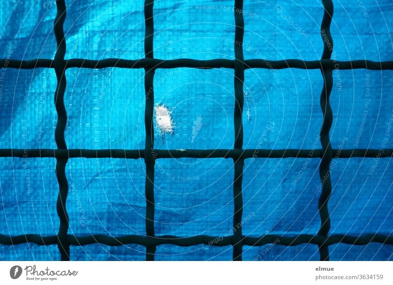 alter Gitterzaun aus Metall, hinterlegt mit blauer, derber Plastikfolie als Sichtschutz, die einen Riss als Guckloch hat Metallzaun PVC-Plane Grenze Schutz