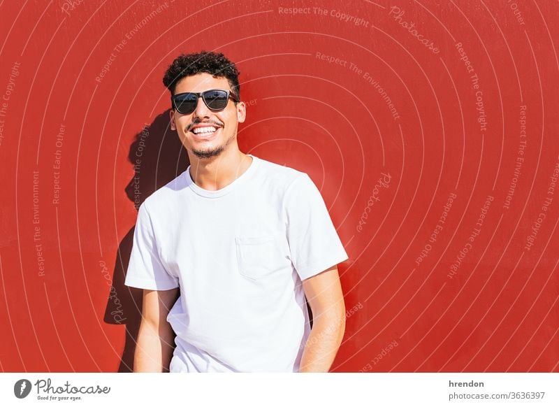 Porträt eines jungen Mannes mit Sonnenbrille, der auf einer roten Wand lächelt Lächeln männlich Typ Lifestyle Glück attraktiv Fröhlichkeit posierend Stil