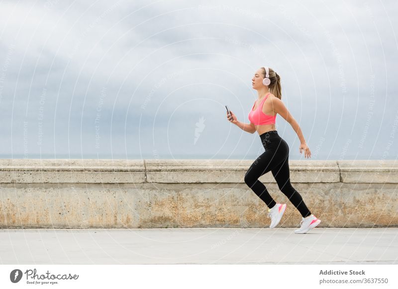 Jogger in Headset einbauen, der mit Smartphone auf der Böschung läuft laufen Kopfhörer Sport Training Sportbekleidung Stauanlage Himmel wolkig schlank Aktivität