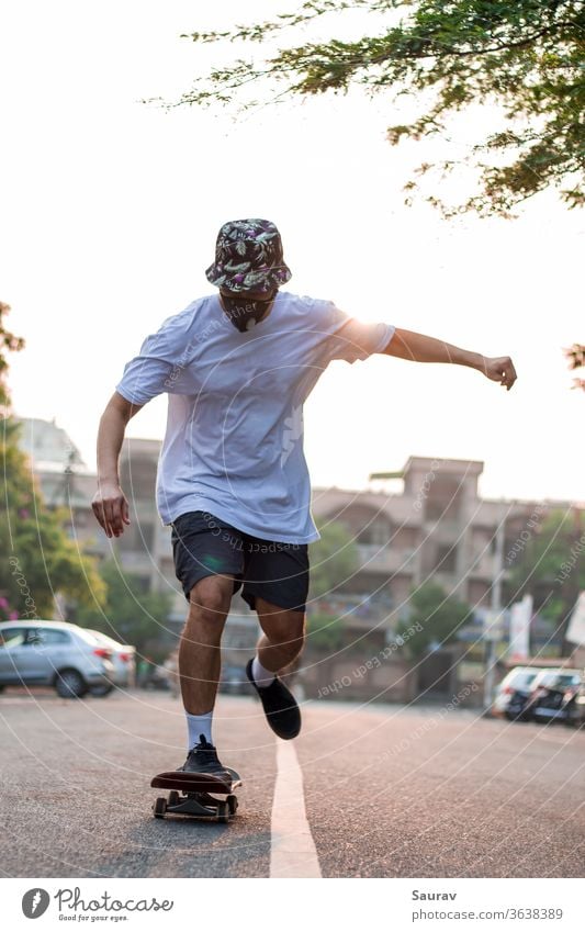Frontansicht eines jungen Erwachsenen, der auf einer leeren Straße Skateboard fährt, während er bei Sonnenaufgang eine Schutzmaske trägt. Sommer Mundschutz