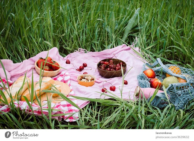 Abfallfreies Sommerpicknick mit Kirschen in den hölzernen Kokosnussschalen, frischem Brot und einer Glasflasche Saft oder Smoothie auf rosa Decke, Flatlay Äpfel