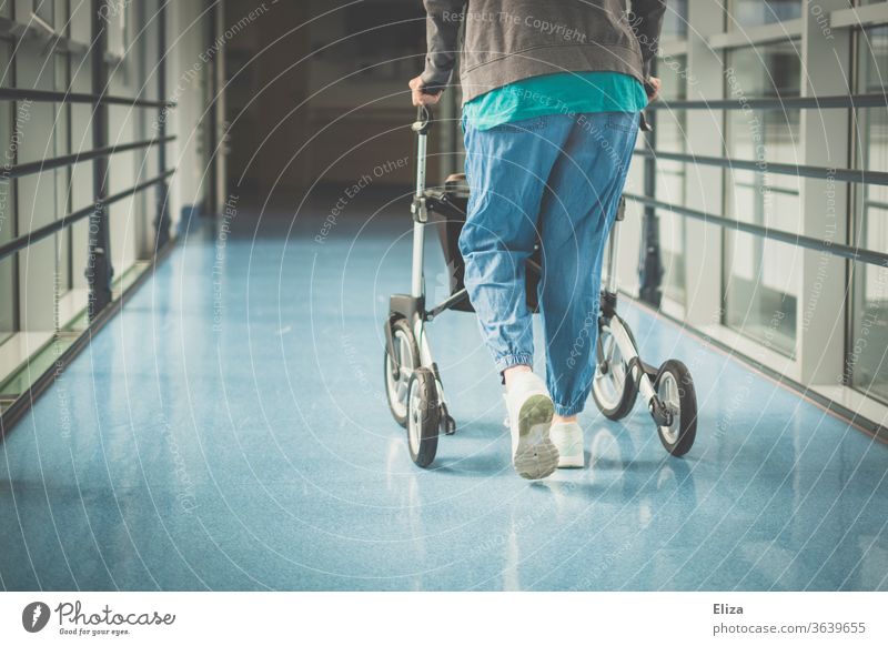 Eine Frau mit Gehbehinderung läuft mit einem Rollator einen Gang im Krankenhaus entlang Behinderung krank alt Gesundheitswesen Mobilität Medizin Pflegeheim