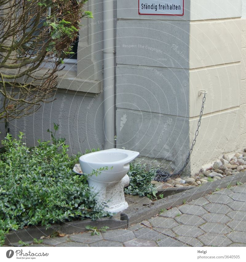 freihalten - ausgediente Toilettenschüssel steht vor einem Haus, an der Wand ein Schild "Ständig freihalten" Klo Klosett Einfahrt Vorgarten Strauch Kette