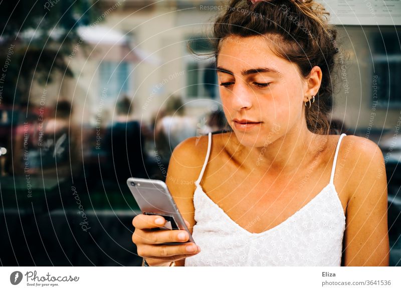 Junge Frau sitzt draußen in einem Café und sieht in ihr Smartphone Sonne Handy lesen beschäftigt junge Frau Telefon Mädchen konzentriert Lifestyle Sommer