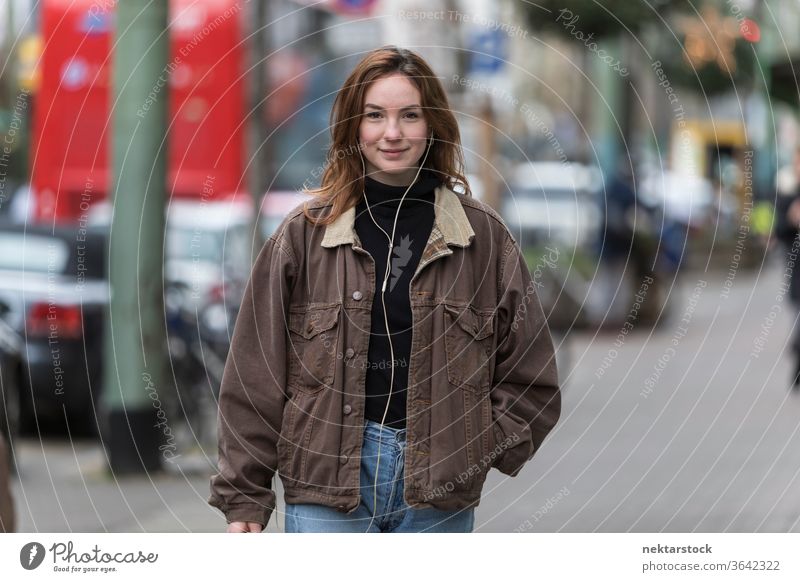 Fröhliche junge Frau hört Kopfhörer auf dem Bürgersteig kaukasische Ethnizität Audio Musik hören Straße Freizeitkleidung Lächeln Hände in den Taschen
