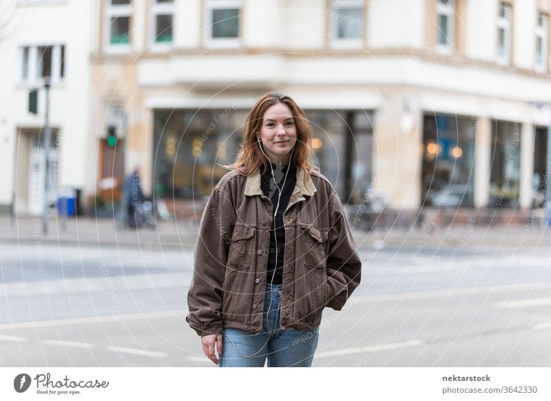 Junge Frau mit echtem Lächeln steht auf der Straße in der Stadt kaukasische Ethnizität Kopfhörer Audio Musik hören Bürgersteig Freizeitkleidung