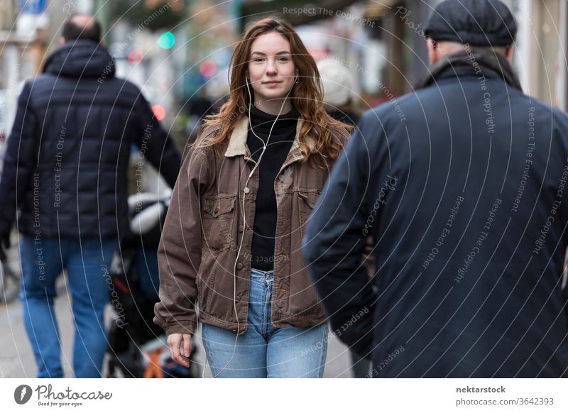 Junge Frau in Jeans und braunem Mantel geht unter Fußgängern kaukasische Ethnizität Kopfhörer Audio Musik hören Bürgersteig Straße Freizeitkleidung braune Haare