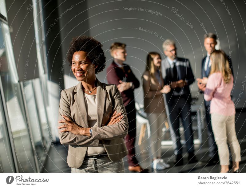 Professionelle schwarze Frau, die mit verschränkten Armen und selbstbewusstem Gesichtsausdruck im Büro steht, während andere Arbeitnehmer im Hintergrund eine Besprechung abhalten