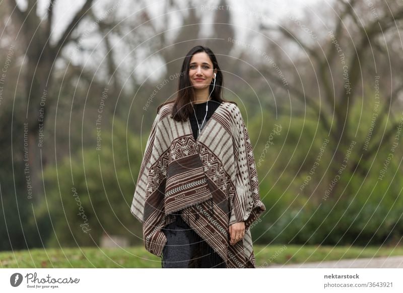 Junge Frau mit gebräunter Hautfarbe, die im Park einen Poncho trägt und lächelt. Ethnie der Indianer nahöstliche Ethnie Kopfhörer mittlere Aufnahme Hörbuch