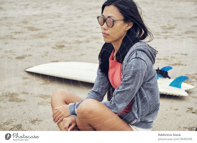 Ruhige Asiatin sitzt am Strand in der Nähe eines Surfbretts Frau Surfer sandig ruhen nachdenklich lässig Sonnenbrille Seeküste sich[Akk] entspannen sitzen jung